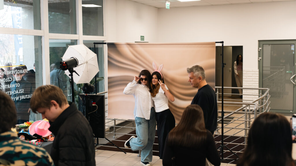 Dwie młode kobiety pozują do zdjęcia na tle fotograficznym, jedna w okularach przeciwsłonecznych, druga w opasce z króliczymi uszami; obok ustawione jest studyjne oświetlenie, a w tle widać innych uczestników wydarzenia.