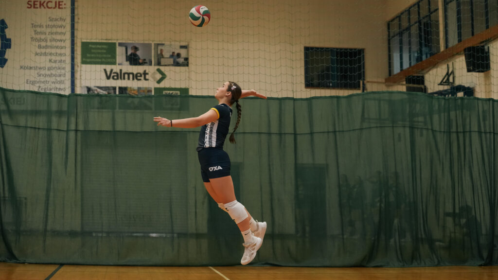 Zawodniczka AZS Humanitas Sosnowiec wykonuje wysoki skok do zagrywki w hali sportowej, z piłką uniesioną nad głową.