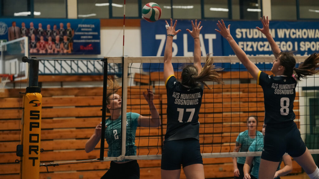 Dwie siatkarki AZS Humanitas Sosnowiec blokują atak zawodniczki drużyny przeciwnej przy siatce podczas meczu na hali sportowej.