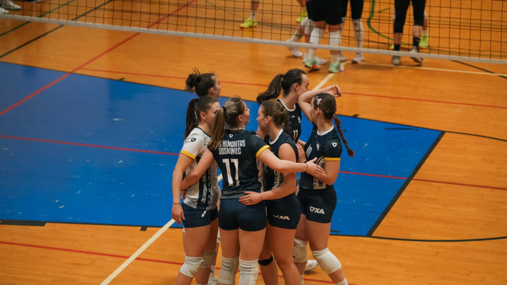 Zawodniczki drużyny siatkarskiej AZS Humanitas Sosnowiec stoją w ciasnym kręgu na boisku, motywując się i omawiając akcję podczas meczu.