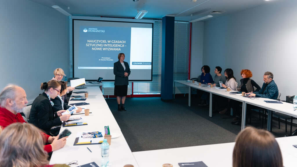 Sala szkoleniowa z uczestnikami siedzącymi przy stołach ułożonych w kształt litery U. Na środku, przed ekranem z prezentacją stoi prowadząca, ubrana z biznesowy strój. Uczestnicy przeglądają materiały i słuchają wystąpienia.