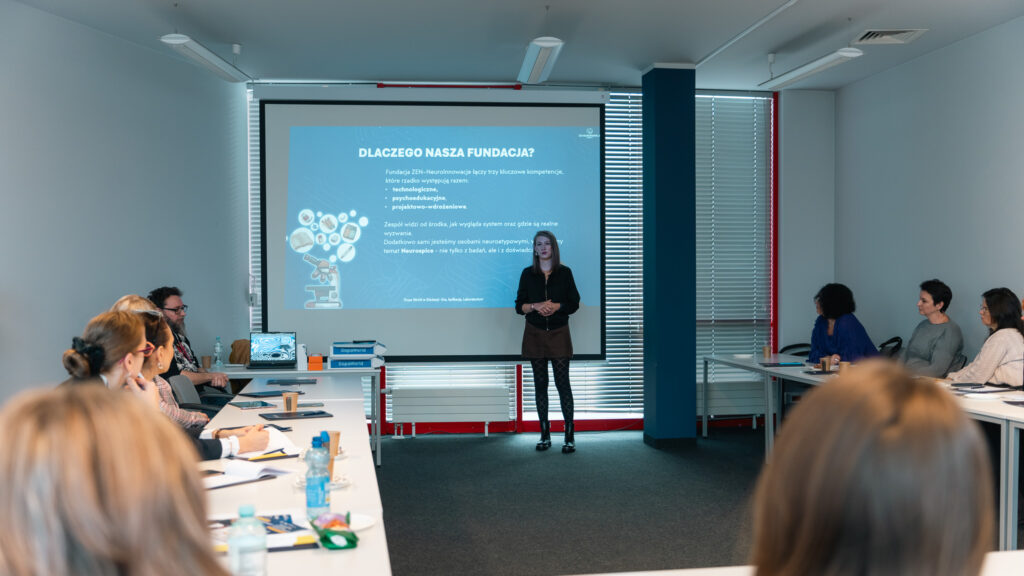 Prelegentka stojąca przed ekranem z prezentacją, prowadząca wykład dla grupy osób - uczestników Brunchu oświatowego Akademii Humanitas - siedzących przy stołach w sali konferencyjnej.