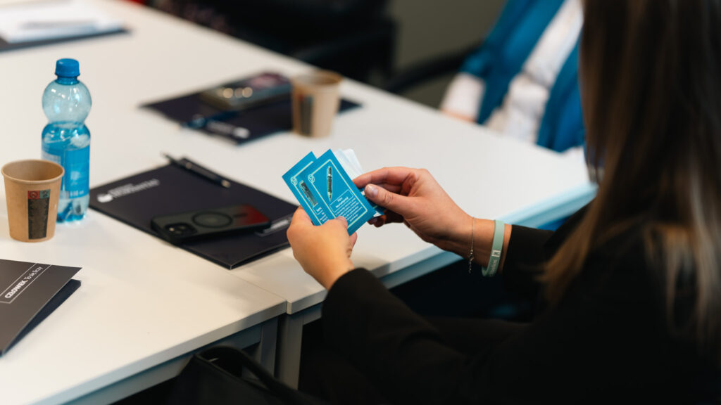 Osoba siedząca przy biurku trzyma w dłoniach kilka niebieskich kart informacyjnych. Na stole przed nią leżą notes, telefon, długopis, papierowy kubek oraz butelka wody. W tle widać fragment drugiej osoby oraz biały blat stołu.