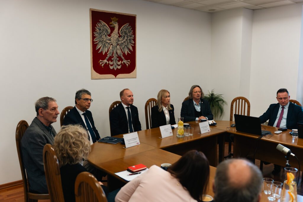 Grupa osób siedzi przy dużym, drewnianym stole podczas formalnego spotkania. Na ścianie za nimi widnieje godło Polski – biały orzeł na czerwonym tle. Uczestnicy mają przed sobą notesy, dokumenty i karafkę z wodą i cytryną. Wszyscy są elegancko ubrani i uważnie słuchają przebiegu spotkania.