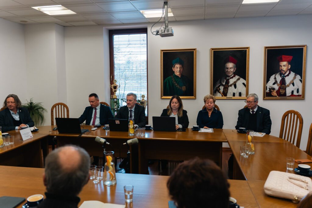 Grupa osób siedzi przy dużym drewnianym stole konferencyjnym w sali uczelni; przed nimi stoją laptopy, mikrofony i dzbanki z wodą. Na ścianie za uczestnikami wiszą trzy portrety rektorów w tradycyjnych togach.