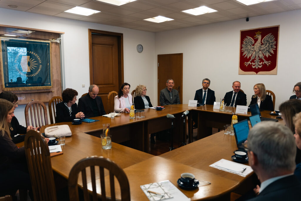 Grupa osób siedzi wokół dużego, drewnianego stołu konferencyjnego podczas oficjalnego spotkania. Na ścianie po prawej stronie widoczny jest herb Polski z białym orłem. Po lewej stronie znajduje się sztandar Akademii Humanitas. Uczestnicy mają przed sobą laptopy, dokumenty i szklanki z wodą, a na środku stołu stoją karafki z cytryną.