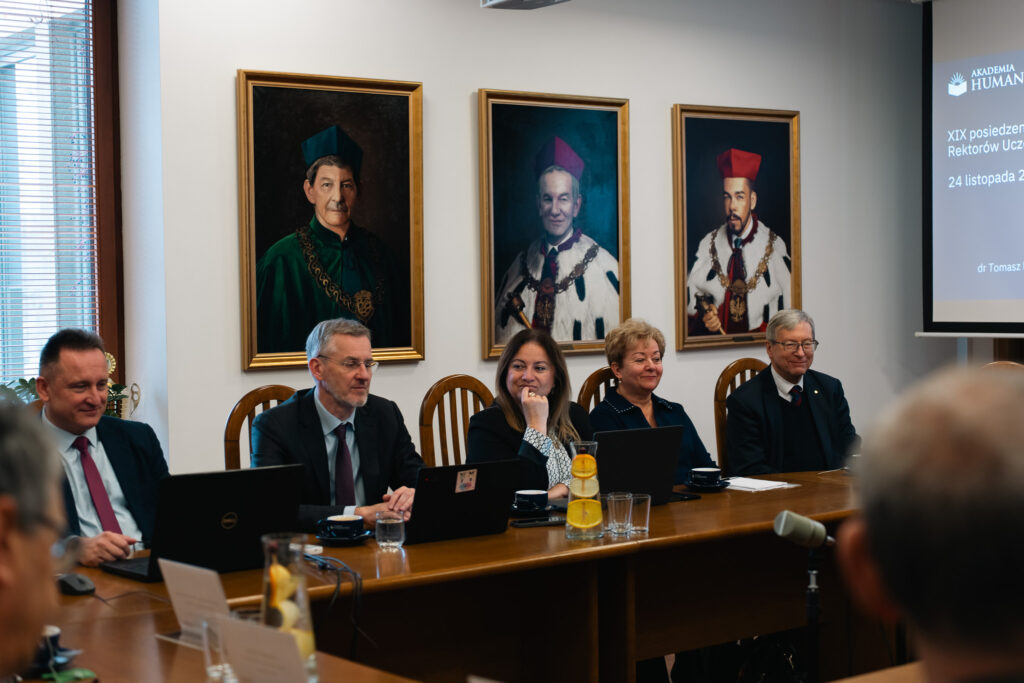 Grupa pięciu osób siedzi przy długim stole podczas oficjalnego posiedzenia. Przed nimi stoją laptopy, szklanki i karafka z wodą i cytrusami. W tle wiszą trzy portrety rektorów w historycznych togach. Po prawej stronie widać fragment ekranu z prezentacją.