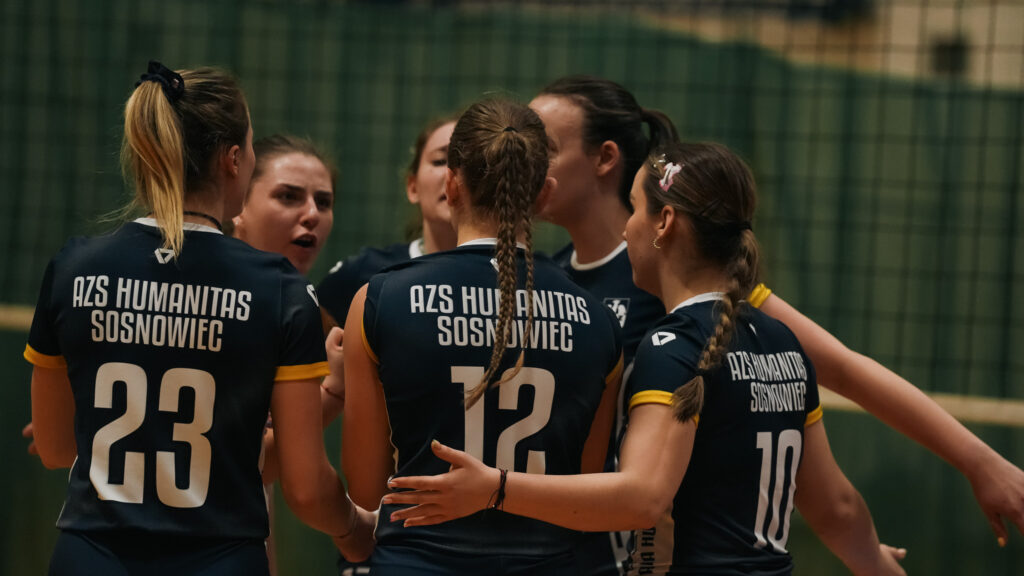 Grupa siatkarek AZS Humanitas Sosnowiec w granatowych strojach stoi w bliskim kręgu, obejmując się i motywując przed kolejną akcją podczas meczu.