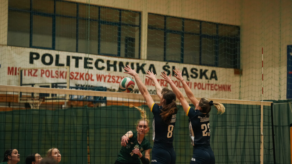 Siatkarka w zielonym stroju atakuje piłkę ponad siatką, podczas gdy dwie zawodniczki z drużyny Akademii Humanitas w granatowych koszulkach z numerami 8 i 23 próbują ją zablokować.