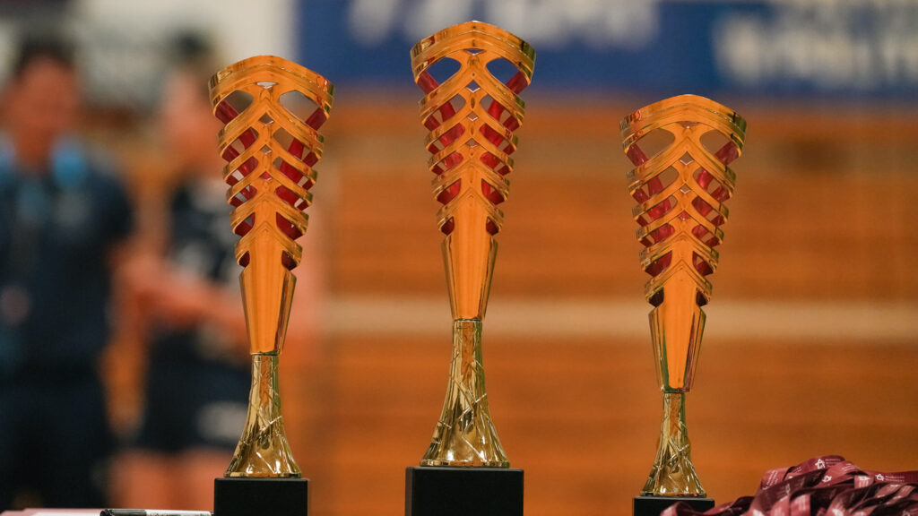 Trzy złote puchary o ażurowym, spiralnym wzorze stoją obok siebie na stole w hali sportowej; na pierwszym planie widać także fragment wstążek od medali.