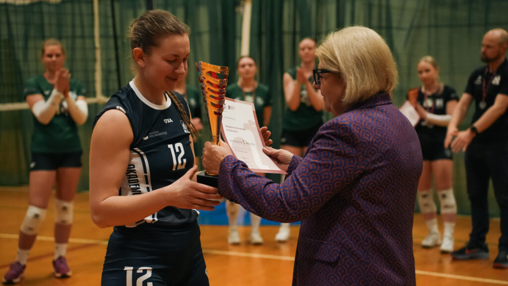 Zawodniczka siatkówki w granatowym stroju z numerem 12 odbiera puchar i dyplom za zajęcie pierwszego miejsca od kobiety w okularach podczas ceremonii na hali sportowej; w tle widać innych zawodników bijących brawo.