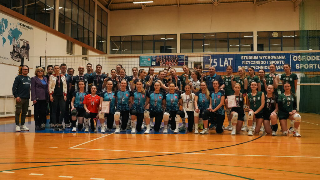 Grupowe zdjęcie trzech drużyn siatkarskich wraz z trenerami i organizatorami, wykonane w hali sportowej po zakończeniu zawodów; zawodniczki prezentują medale, puchary i dyplomy, stojąc i klęcząc przed siatką.