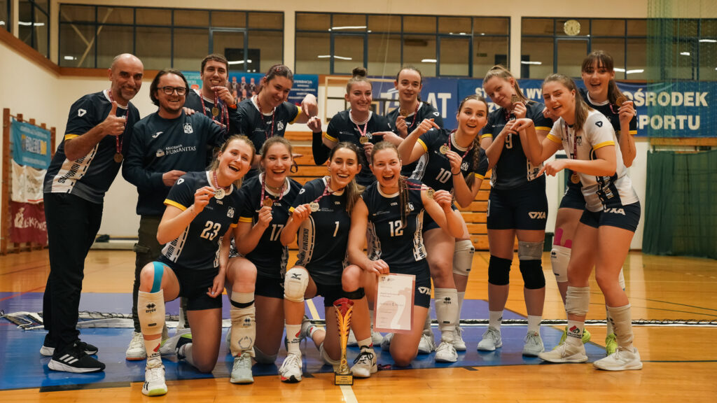 Grupa siatkarek wraz z trenerami pozuje w hali sportowej, uśmiechając się i prezentując zdobyte medale, puchar oraz dyplom po udanym turnieju.