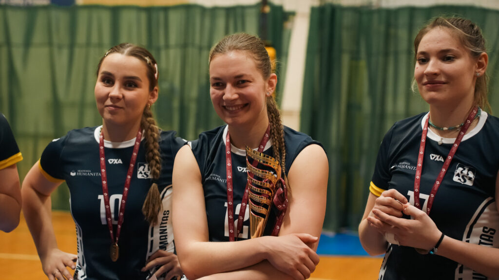 Trzy siatkarki w strojach Akademii Humanitas stoją obok siebie po zakończonym meczu; zawodniczka pośrodku uśmiecha się i trzyma złoty puchar, a wszystkie mają na szyjach medale.