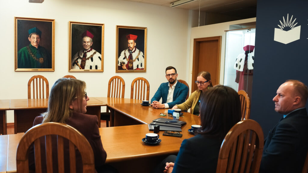 Spotkanie przy stole w Akademii Humanitas, w tle portrety rektorów i insygnia akademickie.