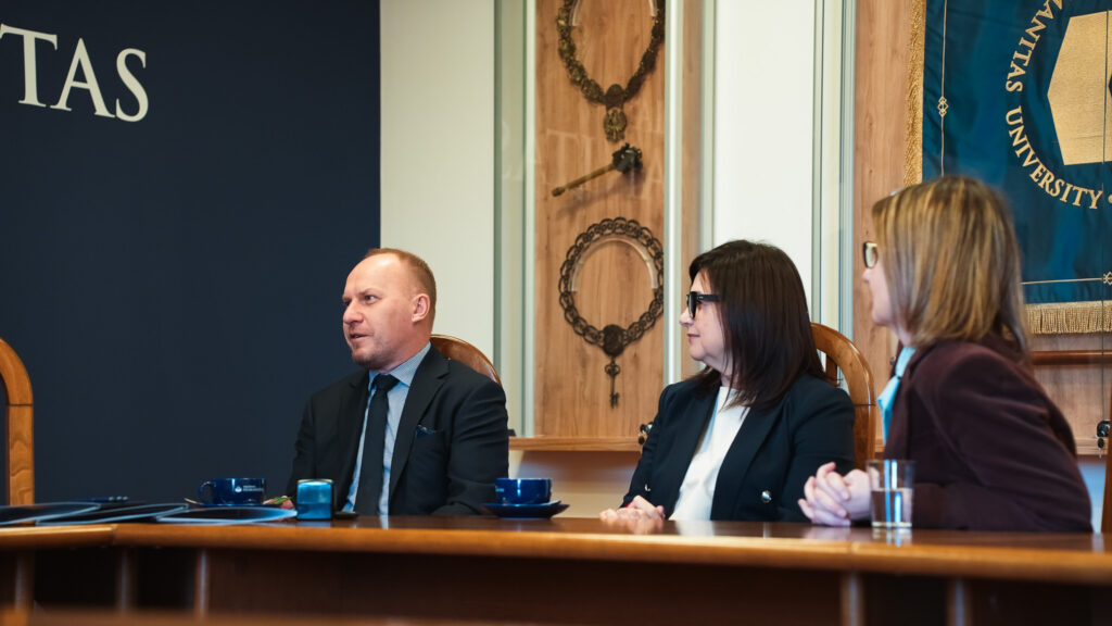 Trzy osoby siedzące przy stole podczas spotkania w Akademii Humanitas, w tle insygnia akademickie i fragment sztandaru uczelni.