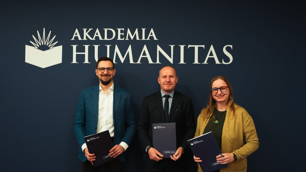 Trzy osoby stojące przed logo Akademii Humanitas, trzymające teczki uczelni i uśmiechające się do kamery.