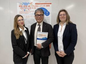 Trzy osoby w formalnych strojach stojące przed plakatem konferencji naukowej o edukacji i dobrostanie w Japonii.