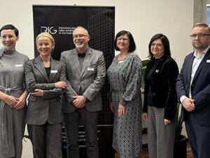 Grupa sześciu osób stoi rzędem na tle roll-upu Regionalnej Izby Gospodarczej w Katowicach. Uczestnicy uśmiechają się do kamery, ubrani w formalne i półformalne stroje.