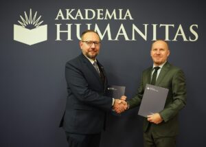 Dwóch mężczyzn w garniturach podaje sobie dłonie w geście powitania lub zawarcia porozumienia. Obaj trzymają teczki z logo Akademii Humanitas. Za nimi znajduje się granatowe tło z dużym białym logotypem i napisem ‘Akademia Humanitas’.