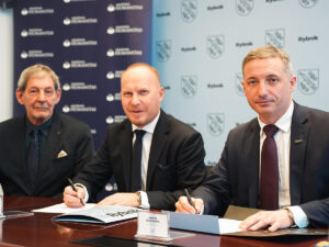 Trzech mężczyzn w garniturach siedzi przy stole i podpisuje dokumenty podczas oficjalnego spotkania, na tle plansz z logotypami instytucji.