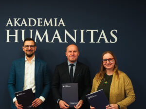 Trzy osoby stojące przed logo Akademii Humanitas, trzymające teczki uczelni i uśmiechające się do kamery.