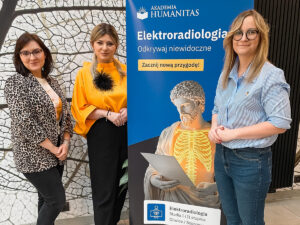 Trzy kobiety stoją obok plakatu promującego kierunek Elektroradiologia na Akademii Humanitas z hasłem „Odkrywaj niewidoczne”.