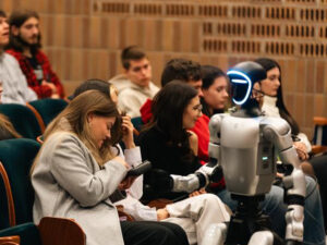 obot humanoidalny siedzi wśród studentów na widowni podczas wydarzenia akademickiego; studenci rozmawiają i sterują robotem.