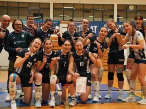 Grupa siatkarek wraz z trenerami pozuje w hali sportowej, uśmiechając się i prezentując zdobyte medale, puchar oraz dyplom po udanym turnieju.
