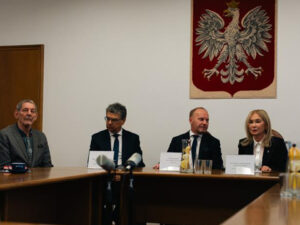 Cztery osoby siedzą przy stole konferencyjnym podczas oficjalnego spotkania; przed nimi stoją tabliczki z nazwiskami, mikrofony oraz dzbanek z wodą i cytryną. Na ścianie w tle widnieje godło Polski.