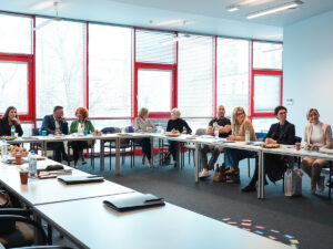 Grupa osób siedzących przy stołach ustawionych w podkowę podczas spotkania lub warsztatów w jasnej sali z dużymi oknami.