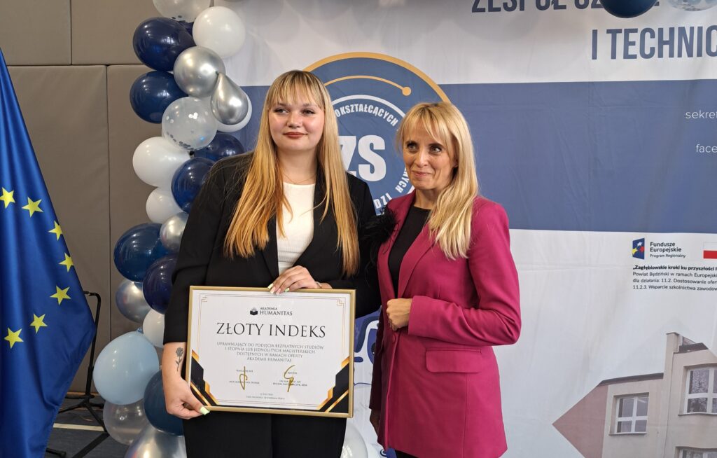 Laureatka Złotego Indeksu pozuje z dyplomem obok przedstawicielki Akademii Humanitas; w tle widoczne są balony w biało-granatowej kolorystyce, flaga Unii Europejskiej oraz baner szkoły.