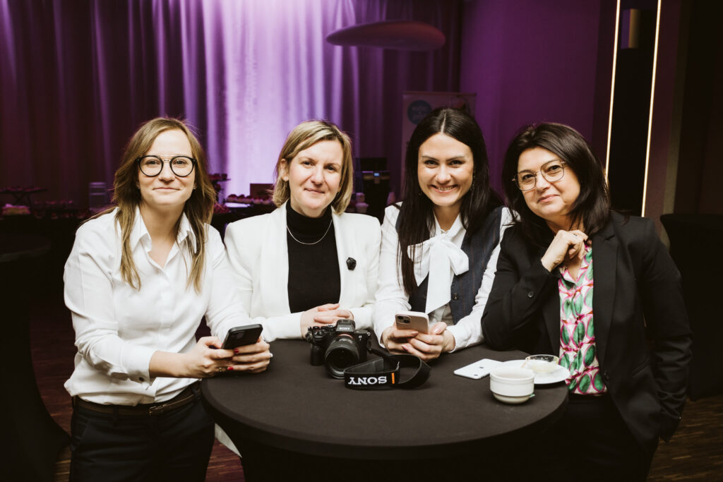 Cztery kobiety stoją przy wysokim stoliku podczas spotkania networkingowego; uśmiechają się do aparatu, trzymają telefony, a na stole leży aparat fotograficzny Sony.