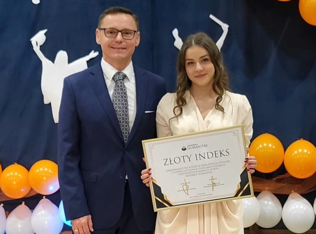 Uśmiechnięta uczennica w jasnej sukience trzyma w ramkach Złoty Indeks Akademii Humanitas, stojąc obok elegancko ubranego mężczyzny na tle dekoracji z balonów i sylwetek absolwentów.