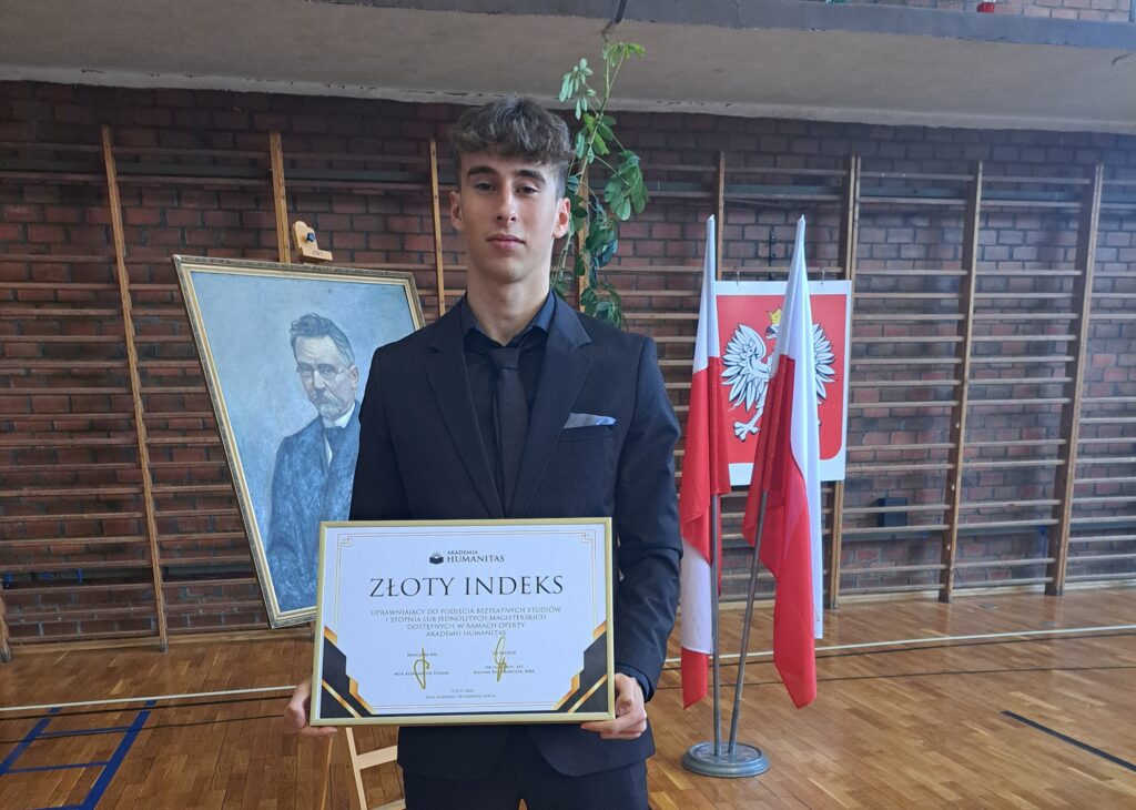 Uczeń w eleganckim garniturze trzyma Złoty Indeks Akademii Humanitas; za nim widoczne są polskie flagi oraz portret na sztaludze ustawione w sali gimnastycznej.