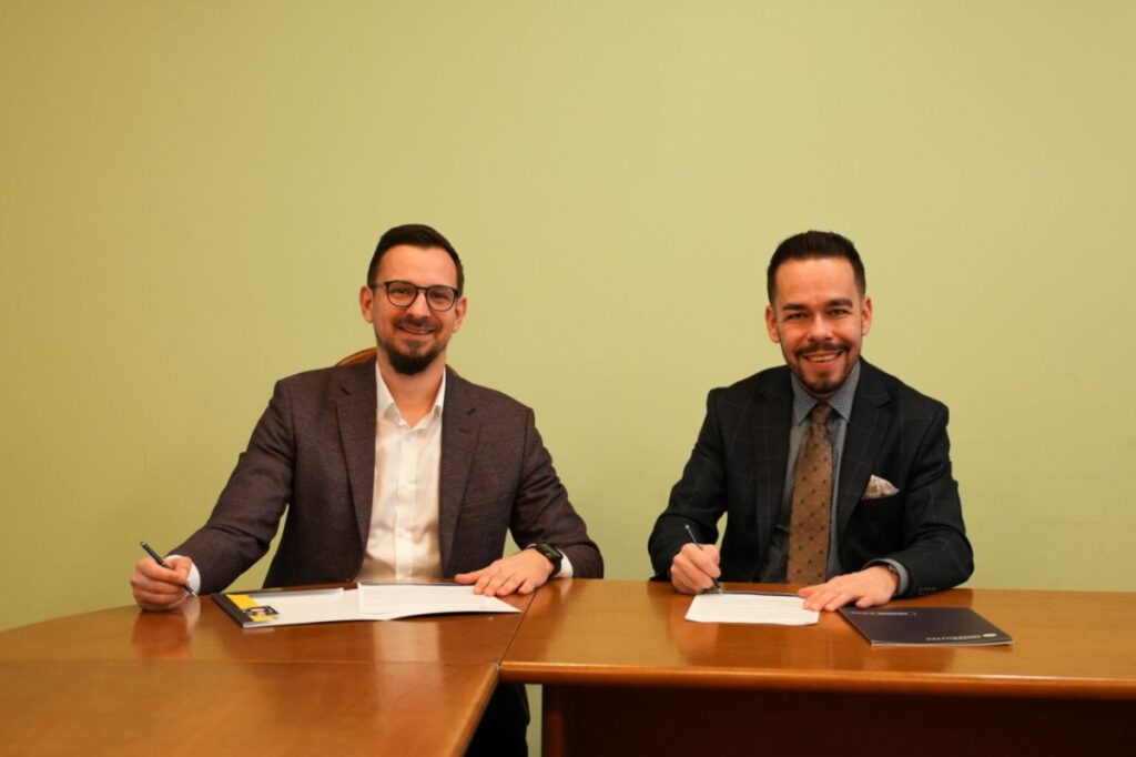 Dwóch mężczyzn w eleganckich marynarkach siedzi przy stole i podpisuje dokumenty, uśmiechając się w stronę aparatu; na stole leżą papiery i teczka z logo Humanitas, w tle jasna, jednolita ściana.