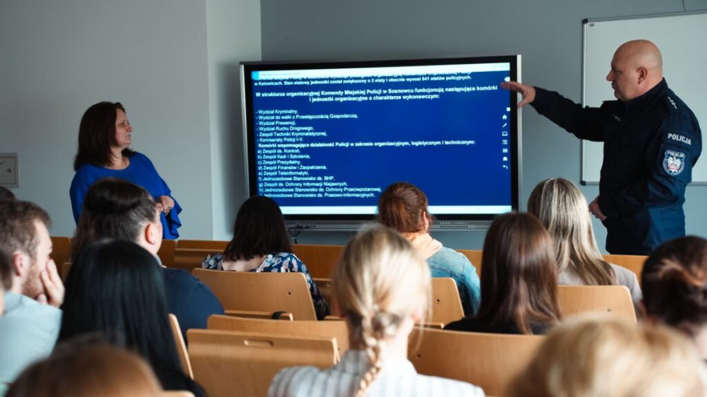 Policjant prowadzi prezentację przed grupą słuchaczy w sali wykładowej, wskazując na slajd wyświetlany na ekranie, obok stojącej prowadzącej.