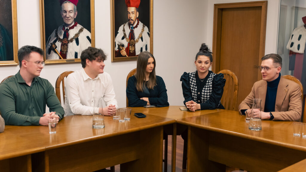 rupa młodych dorosłych rozmawia w sali konferencyjnej, w tle portrety rektorów w ceremonialnych strojach. Uczestnicy siedzą naprzeciw siebie, prowadząc dyskusję w formalnym, akademickim otoczeniu.