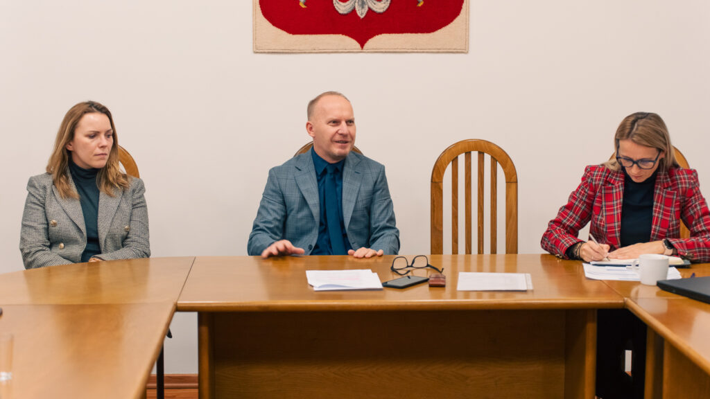 Spotkanie przy drewnianym stole – trzy osoby siedzą w sali konferencyjnej, mężczyzna pośrodku prowadzi rozmowę, a kobieta po prawej sporządza notatki. Formalne, urzędowe wnętrze podkreśla charakter roboczego spotkania.