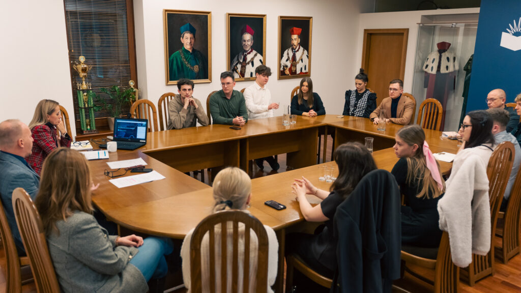 Spotkanie przy dużym, drewnianym stole – kilkanaście osób uczestniczy w dyskusji w sali konferencyjnej, w tle portrety rektorów i symbole uczelni. Atmosfera formalnej narady akademickiej.