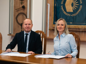 Na zdjęciu dwie osoby siedzą przy stole i podpisują dokumenty; mężczyzna w garniturze po lewej oraz kobieta w jasnoniebieskiej koszuli po prawej, oboje uśmiechnięci. W tle widoczne są symbole uczelni, w tym niebiesko-złoty sztandar Akademii Humanitas.