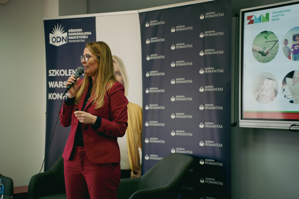 Kobieta w bordowym garniturze przemawia do publiczności, trzymając mikrofon w sali konferencyjnej. W tle widoczne są roll-upy Akademii Humanitas oraz ekran z prezentacją.