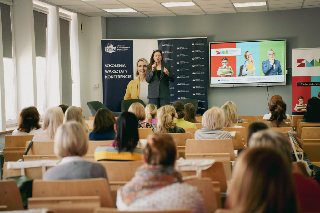 Prelekcja podczas wydarzenia Akademii Humanitas – kobieta w garniturze przemawia do uczestników zgromadzonych w sali wykładowej; w tle roll-upy „Szkolenia, warsztaty, konferencje” oraz ekran z prezentacją.