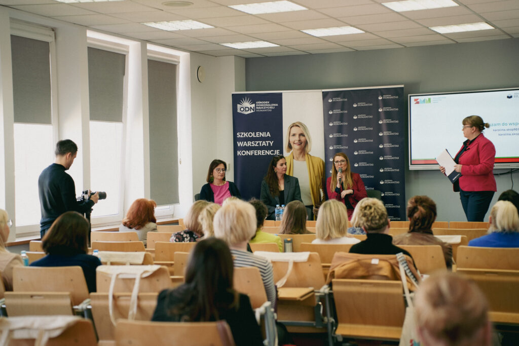 Panel dyskusyjny podczas wydarzenia Akademii Humanitas – trzy prelegentki siedzą przy stole i rozmawiają z publicznością, jedna z nich mówi do mikrofonu; po prawej prowadząca z dokumentami, w tle banery „Szkolenia, warsztaty, konferencje” oraz ekran prezentacji.
