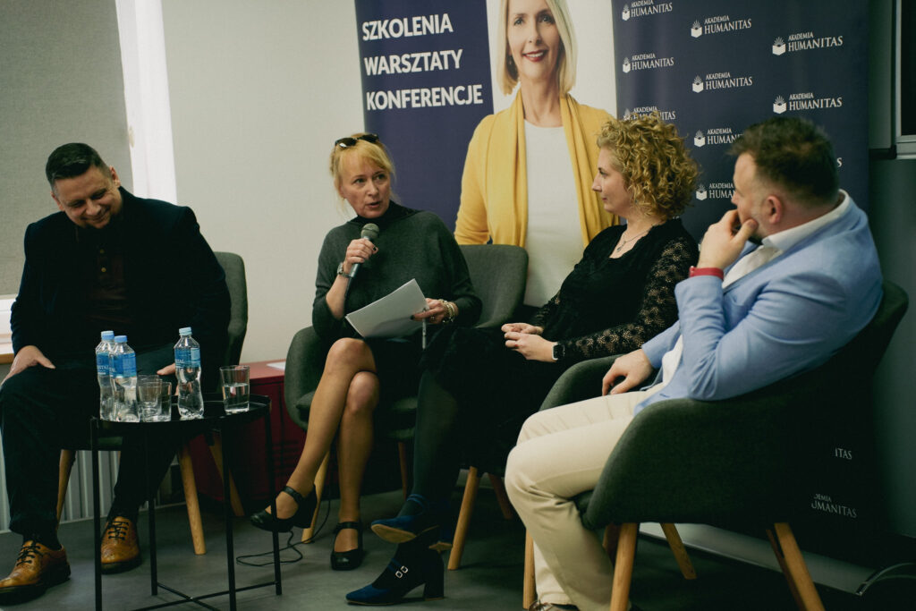 Panel dyskusyjny podczas wydarzenia Akademii Humanitas – cztery osoby siedzą w fotelach, jedna z kobiet mówi do mikrofonu i trzyma notatki; w tle roll-upy z hasłami „Szkolenia, warsztaty, konferencje”.