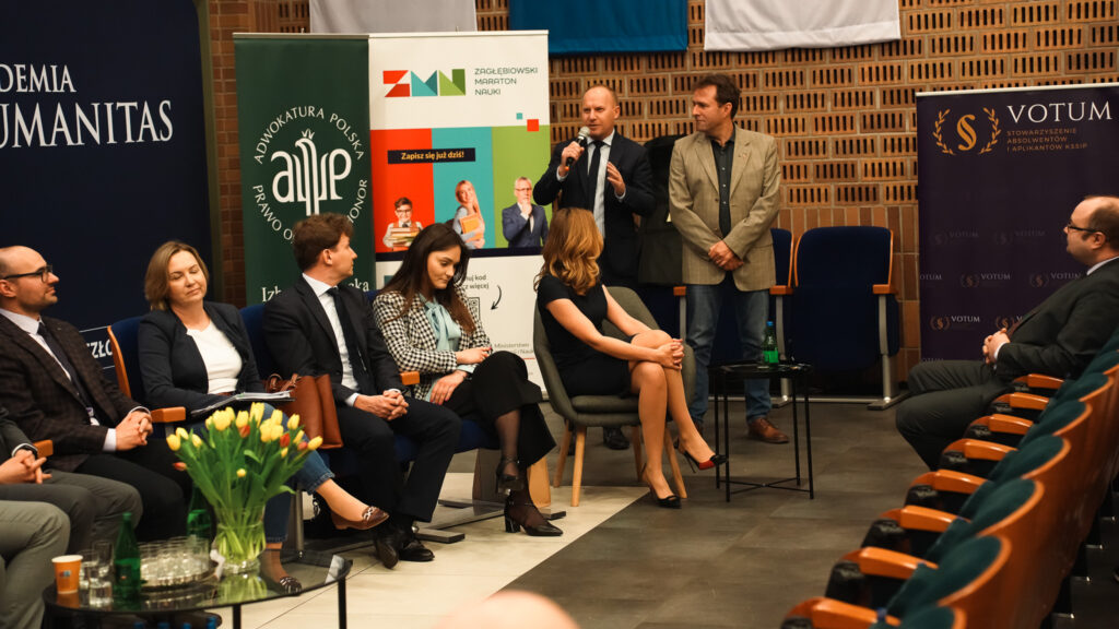 Dyskusja panelowa w Akademii Humanitas – prelegent z mikrofonem przemawia do publiczności, pozostali uczestnicy siedzą na scenie obok banerów partnerów wydarzenia.