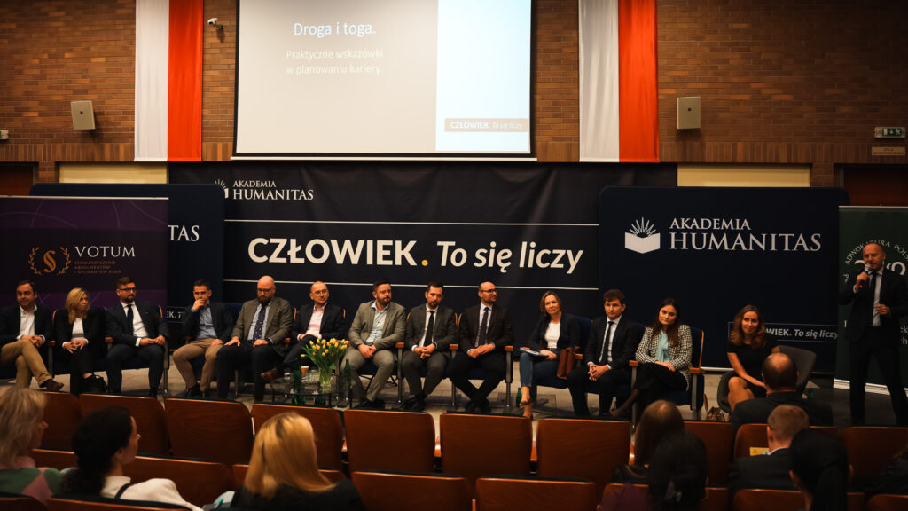 Panel dyskusyjny z udziałem prelegentów siedzących na scenie w auli Akademii Humanitas, przed banerem z hasłem »Człowiek. To się liczy«. Na widowni zgromadzona publiczność, wydarzenie o charakterze akademicko-społecznym.