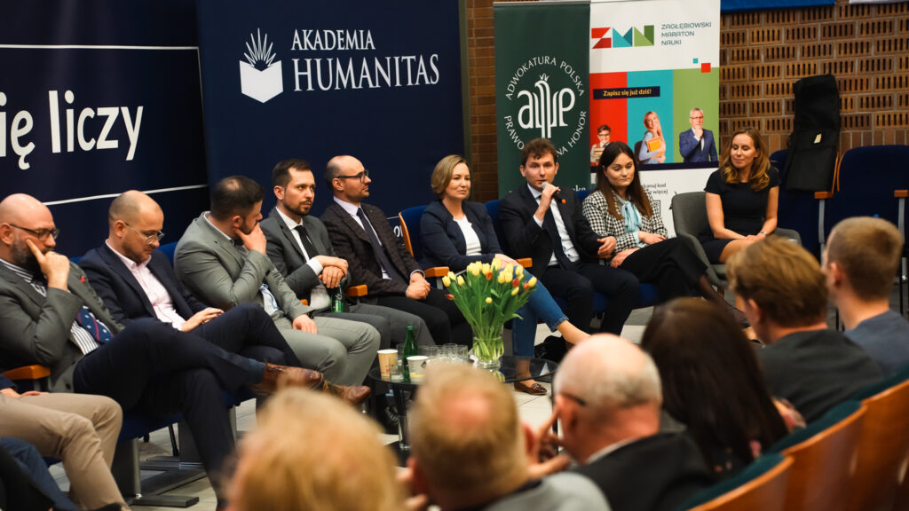 Panel dyskusyjny w Akademii Humanitas – uczestnicy siedzą na scenie, jeden z prelegentów mówi do mikrofonu, a publiczność słucha w auli.
