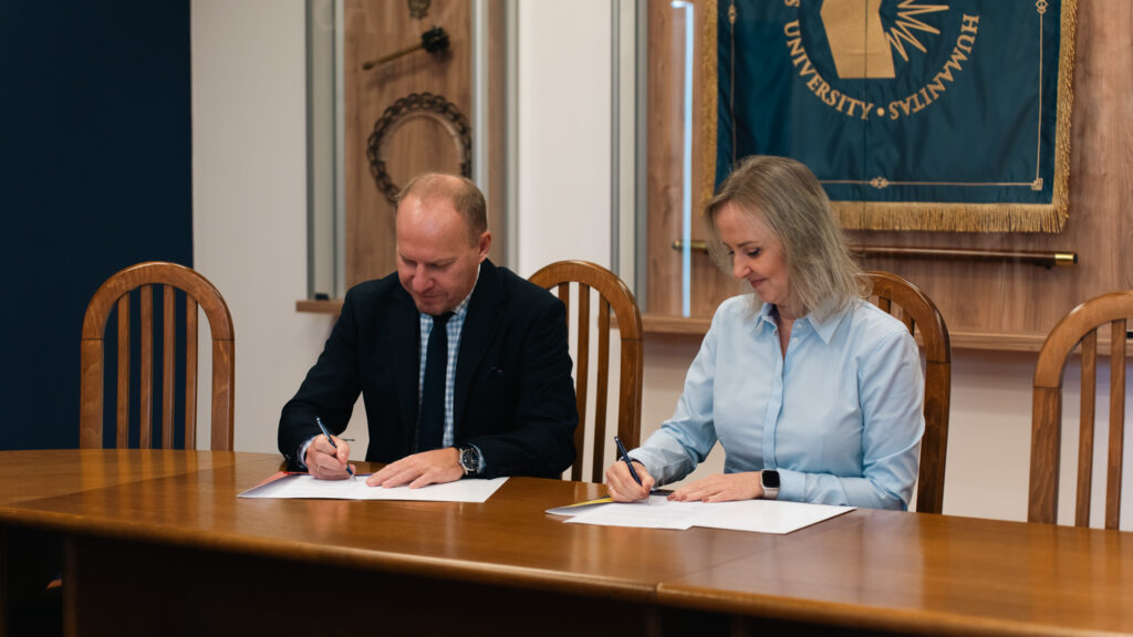 Mężczyzna w garniturze i kobieta w jasnoniebieskiej koszuli siedzą przy drewnianym stole i podpisuje dokumenty; mężczyzna w garniturze pisze po lewej stronie, a kobieta w jasnoniebieskiej koszuli po prawej, w tle widoczne są elementy dekoracyjne oraz sztandar uczelni.