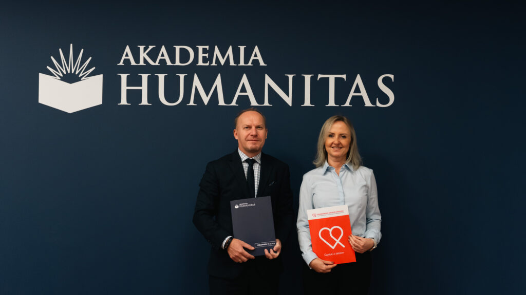 Mężczyzna w garniturze i kobieta w jasnoniebieskiej koszuli stoją obok siebie na tle granatowej ściany z dużym logo Akademii Humanitas, trzymając w rękach teczki – on granatową uczelnianą, ona czerwoną z logo Zagłębiowskiego Centrum Onkologii. 